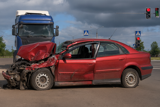 A red sedan with severe front-end damage sits at an intersection in Alabama, in front of a large blue semi-truck, with traffic lights and crosswalk signs visible in the background.