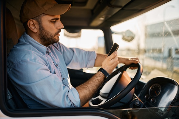 A male truck driver wearing a baseball cap sits in the cab of his truck and looks down at the cell phone he is holding while driving. (503962124)