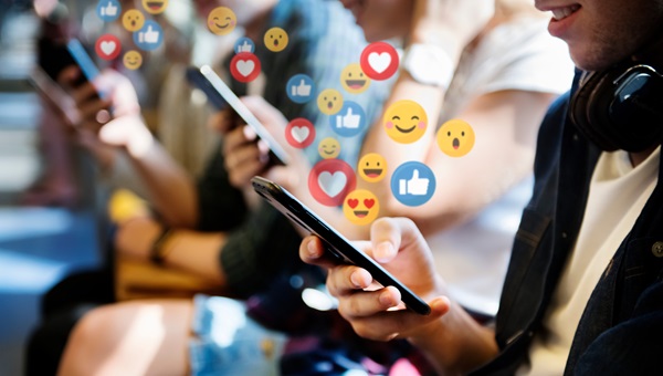 Several teenagers sit in a row using their smartphones as a variety of colorful social media icons, including hearts, thumbs-up, and emojis, float above the screens to illustrate the focus of Alabama social media addiction legal claims.