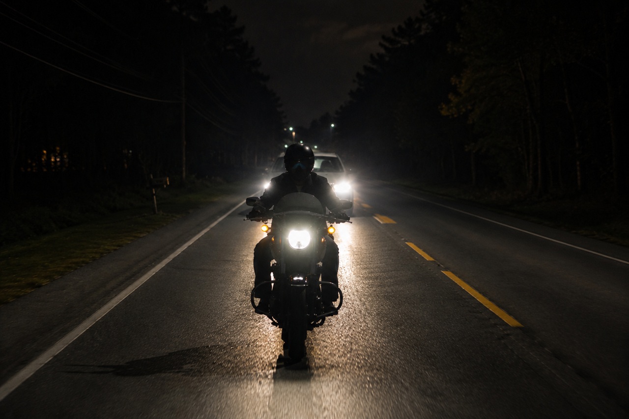A motorcyclist riding on a dark, tree-lined two-lane road at night with a car's bright headlights visible closely behind them, illustrating the limited visibility and rear-end hazards that contribute to nighttime motorcycle accident risks in Alabama.